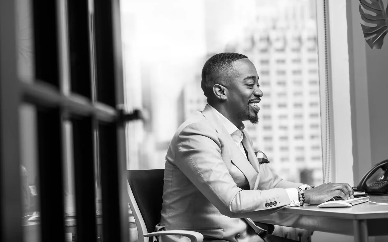 A man working at a laptop by a window with a city view, in black and white