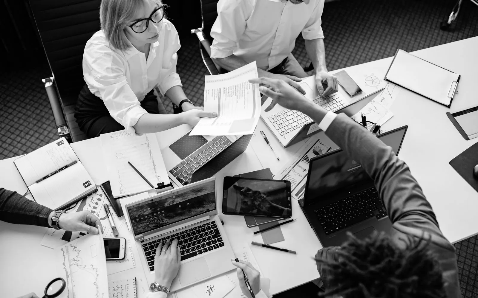 Colleagues working over documents and laptops at a desk, seen from above, in black and white